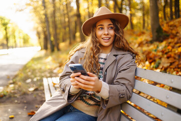 A cheerful woman in a hat and with a phone sits on a wooden bench in an autumn park. A beautiful...