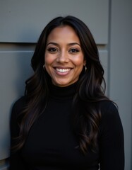 A portrait headshot photo of a professional ceo executive business worker: a black woman with long hair and a warm smile wearing gold earrings and a black turtleneck standing in front of a gray wal