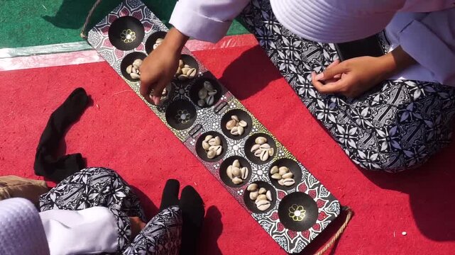 High-angle view of two people playing a traditional mancala-style board game, known as Congklak or Dakon, using cowrie shells