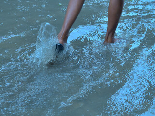 Bare legs walking through water, creating splashes. The water appears clear, reflecting bluish hues. Only the lower legs are visible, with one foot wearing a sandal, indicating motion.