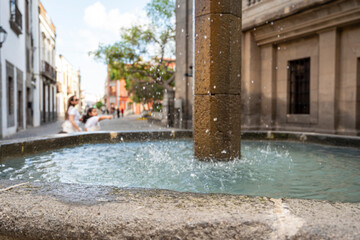 A fountain with water splashing out of it. The water is blue and the fountain is surrounded by a stone wall. There are people walking by and sitting on the sidewalk. The scene is lively and peaceful