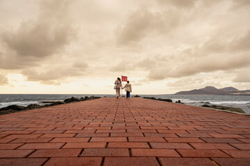Fototapeta premium Mother and daughter walk along a brick pavemen. The sky is cloudy and the water is in the background