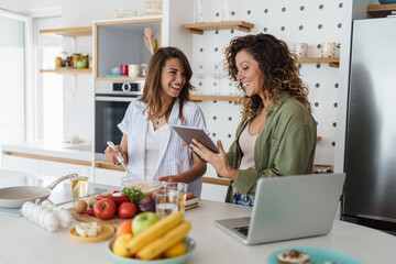 Two Happy Women Cooking in a Bright Modern Kitchen Using Fresh Ingredients and a Tablet
