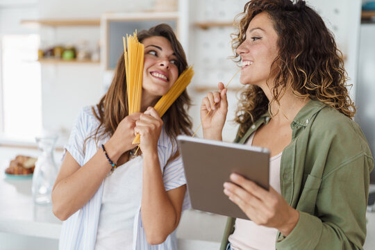 Young Happy Women Cooking Together in a Bright Modern Kitchen Holding Pasta and a Digital Tablet - Powered by Adobe