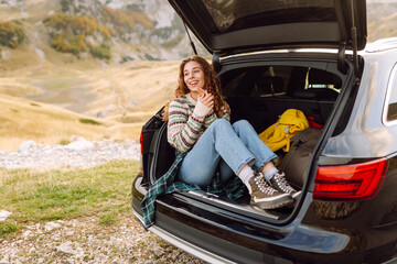 Naklejka premium Smiling woman sitting in the trunk of a car and enjoying the mountain landscape on a sunny day. Beautiful female hiker on a road trip feels freedom. Nature and freedom concept.