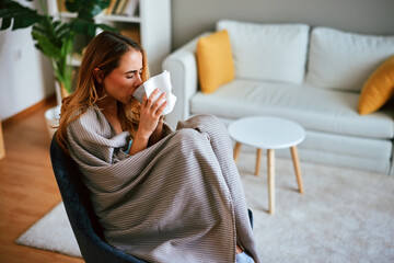 Sick woman drinking hot beverage at home covered with blanket