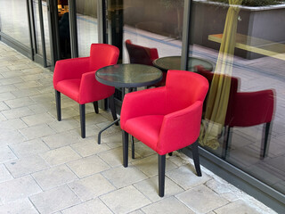 Two red upholstered chairs with a small round table placed on a cafe terrace by glass wall. Hospitality, lifestyle and dining environment reflecting comfort, leisure and modern design.