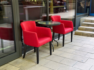 Two red upholstered chairs with a small round table placed on a cafe terrace by glass wall. Hospitality, lifestyle and dining environment reflecting comfort, leisure and modern design.