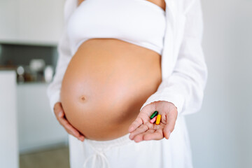 A close-up of a pregnant woman's belly holding colorful vitamins in her hand. A young mother of an...