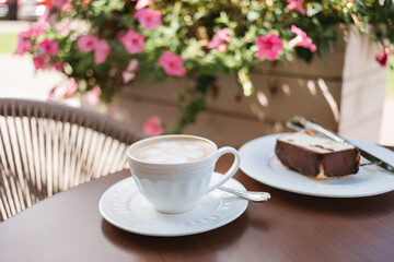 A white cup of cappuccino on a table next to a delicious slice of cake on the terrace of a cafe, decorated with planters full of flowers.