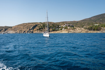 White sailboat sailing in the blue waters of the mediterranean sea, with the coastal town of...