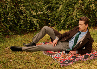 Man in stylish outfit relaxing on a blanket surrounded by greenery.