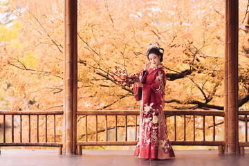 A woman in a red traditional dress poses with flowers among autumn leaves.