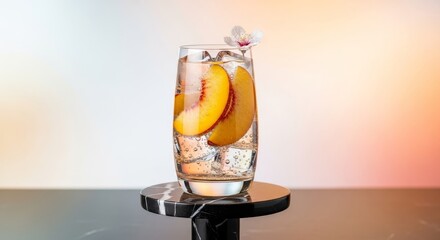 A highball glass with peach slices ice cubes and a flower displayed on a marble stand against a blurred background