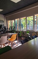 An interior shot of a modern office space or cafe, featuring a grid ceiling, large windows with a view of a sunlit forest, and several colorful chairs around a table.