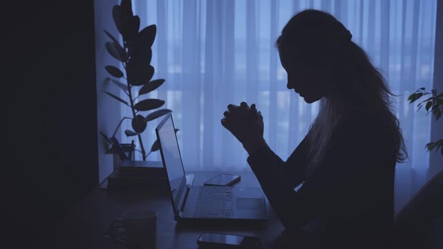 dark unrecognizable silhouette unhappy tired woman working on laptop sitting at desk in black room window light. Sad girl suffers from headache depression fatigue lack of money, Person looking for job