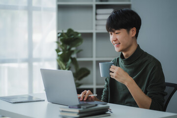 Obraz premium Asian freelancer working remotely using laptop and drinking coffee from mug, sitting at desk in home office