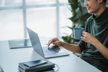 Asian male designer holding a coffee mug while working on his laptop with a stylus pen in his home office