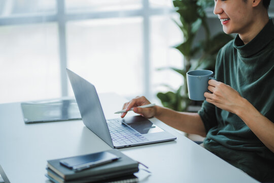 Asian male designer holding a coffee mug while working on his laptop with a stylus pen in his home office - Powered by Adobe