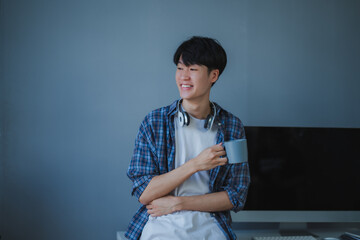 Smiling Asian developer enjoying a coffee break, relaxing at his desk in a modern office while contemplating ideas and inspiration