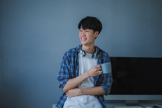 Smiling Asian developer enjoying a coffee break, relaxing at his desk in a modern office while contemplating ideas and inspiration