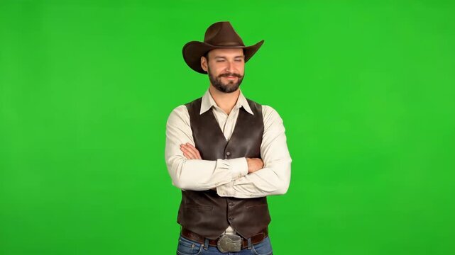 A man in a cowboy hat and vest stands with his arms crossed against a green screen background