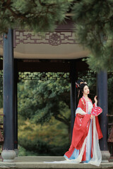 Woman in traditional red and blue hanfu holding a fan in a serene outdoor gazebo Chinese word means...