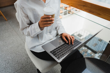 Businesswoman sitting and using laptop while holding takeaway coffee cup, working remotely in...