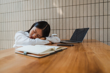 Young asian office worker sleeping at desk with laptop, smartphone, notebook and takeaway coffee on wooden table