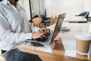 Businesswoman typing on laptop keyboard at glass desk in modern office with takeaway coffee cup