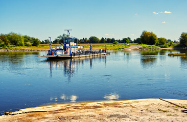 Fototapeta premium Ferry traversing elbe river near sandau, germany, with verdant landscape and azure sky during summer sunlight