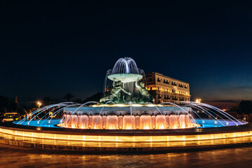 The Triton Fountain illuminated at night, featuring three bronze figures holding a large basin at...