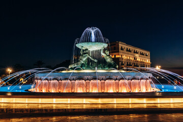 The Triton Fountain illuminated at night, featuring three bronze figures holding a large basin at the entrance to the city of Valletta