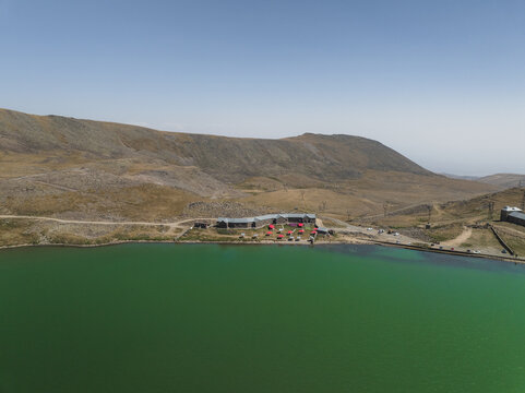 Aerial view of the vivid green lake contrasting sharply with the arid, rocky terrain, framed by distant mountains and a clear blue sky, Lake Kari, Aragatsotn Province, Armenia.