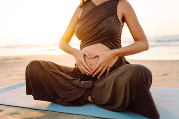 A woman sits on a yoga mat and touches her pregnant belly in the rays of the sunset. Close-up of a young woman practicing yoga and meditation on the seashore. Concept of procreation, health.