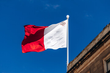 Naklejka premium The national flag of Malta, red and white with the George Cross in the upper hoist corner, flies against a clear blue sky above traditional Maltese limestone architecture