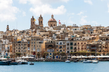 Naklejka premium Birgu, Malta – April 11, 2025: Picturesque Maltese harborfront with traditional limestone buildings, colorful boats, and waterfront cafés, set beneath a bright sky and grand church domes