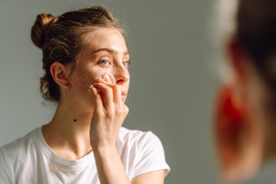 Close up of young woman face wiping face with cotton pad looking in mirror. Cute woman enjoying natural beauty, wiping makeup in cozy room. Beauty, skin care concept.