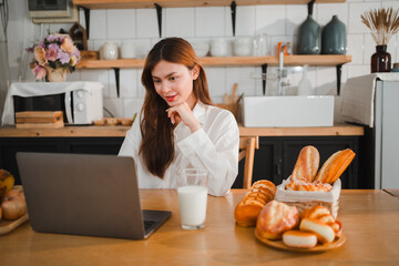 Young woman using laptop in kitchen with pastries and milk