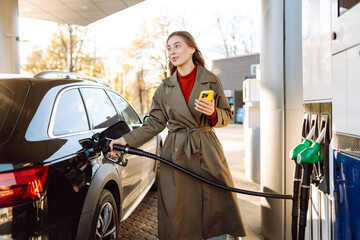 A young woman with a phone fills up her car at a gas station. A woman at a gas station fills up her tank with fuel using an app on her phone. Transport concept, industry.