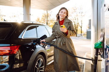 A young woman with a phone fills up her car at a gas station. A woman at a gas station fills up her...