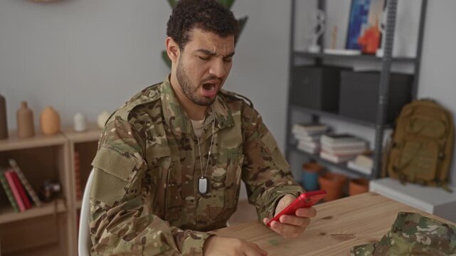 Hispanic man in camouflage uniform holds smartphone and taps screen in building, cringing at message received; stress frustration isolation anxiety.