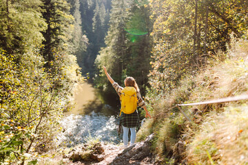 Female hiker with bright backpack walking on pedestrian suspension bridge in mountains. Young woman enjoying mountain scenery and feeling freedom in open air. Travel, freedom concept.