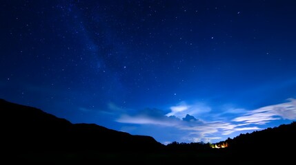A serene night sky filled with stars, illuminated by a soft blue hue, with silhouettes of mountains in the foreground and distant lights from a small settlement