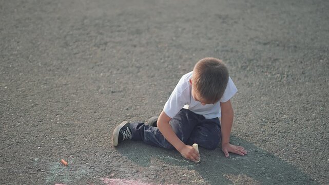 The boy, cheerful child, sits on street with chalk in hand. The kid enjoys outdoor play, creating summer drawing. Street art comes alive as boy, child, and kid make colorful chalk creations together.