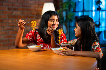 Two Indian sisters eating noodles together on sofa during casual snack time at home