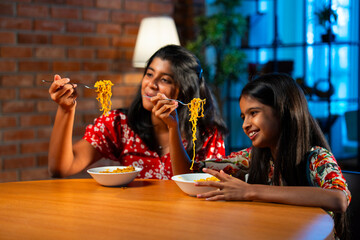 Two Indian sisters eating noodles together on sofa during casual snack time at home