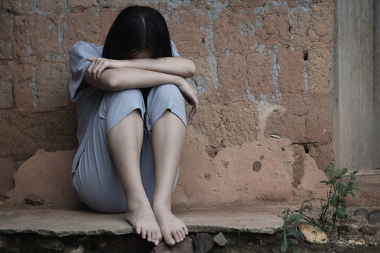 Poor,depressed young girl sits next to an old brick wall her head bowed and cries.