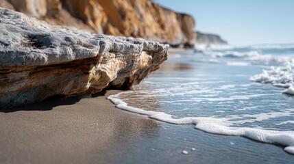 A close-up view of a rocky shoreline, with waves gently lapping the sandy shore, creating a tranquil scene under a bright, clear sky.