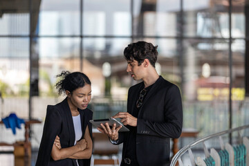 Young business officers in black jacket exchange ideas on business using tablet to provide data and information at a terrace outside of the building. Conversation of two coworkers in casual dress